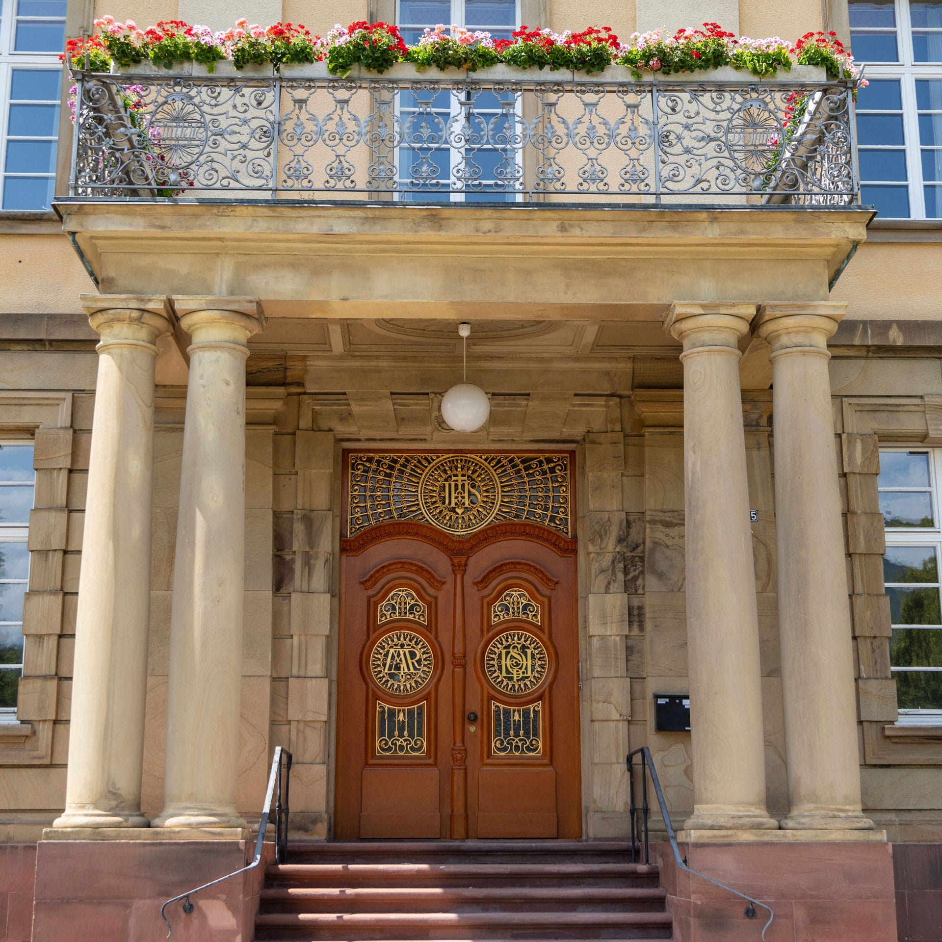 Ein Gebäudeeingang mit zwei kunstvoll verzierten Holztüren, vier Steinsäulen, einem Balkongeländer aus Metall und Blumenkästen auf dem darüber liegenden Balkon – ideal für alle, die die Eleganz des Palais Erlenbad oder der Premium-Apartments Sasbach suchen.